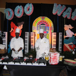 Two chefs standing in front of a table with a Doo Wah Stage and Table Set.