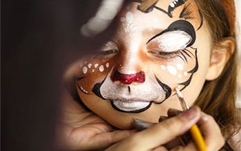 A girl is getting her face painted with a tiger by a Caricature Artist.